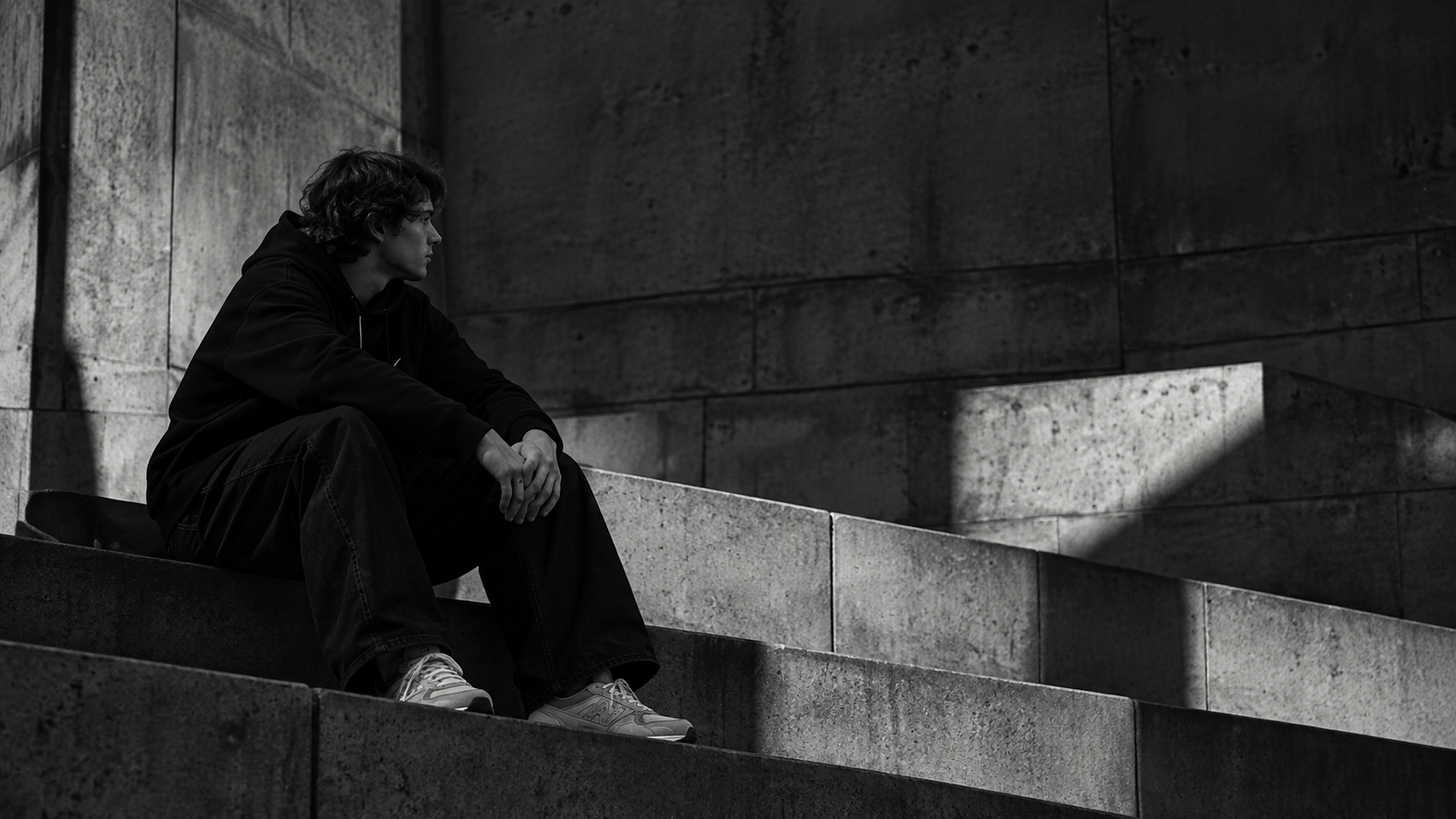Joven sentado en unas escaleras de concreto, a la izquierda de un muro en sombras. Fotografía en blanco y negro.